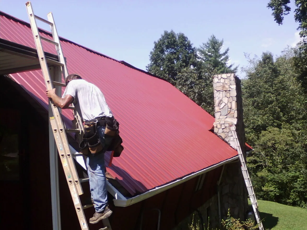 Roofer on a ladder beside a red metal roof and stone chimney — Soffit & Fascia Repair by Roofing Brothers in South Lebanon